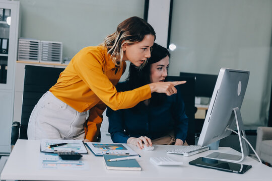 Two businesswomen discussing work using a laptop in a modern office lounge area, showing teamwork and communication
