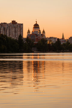 Golden sunset over the Dnipro River at Obolon Embankment in Kyiv, Ukraine, with the Church of the Nativity of Christ and its golden domes reflected in the calm water.