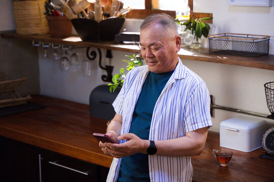 Senior Asian man leaning on wooden countertop in home kitchen holding smartphone and tea cup