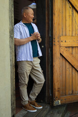 Fototapeta premium Senior Asian man standing on threshold in striped tee, leaning against planked door, clasping hands