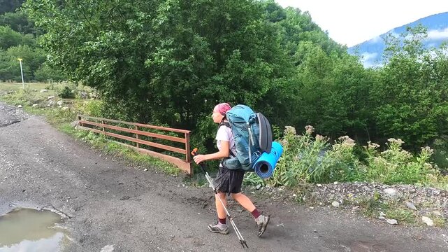 Female backpacker walks muddy trail, side view with loaded pack and trekking poles in Svaneti, Georgia, representing a multiday hiking, equipment transport and self-supported mountain travel