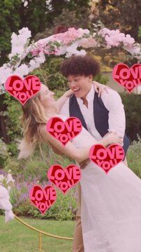 Animation of red love you hearts over happy diverse couple embracing in garden on wedding day