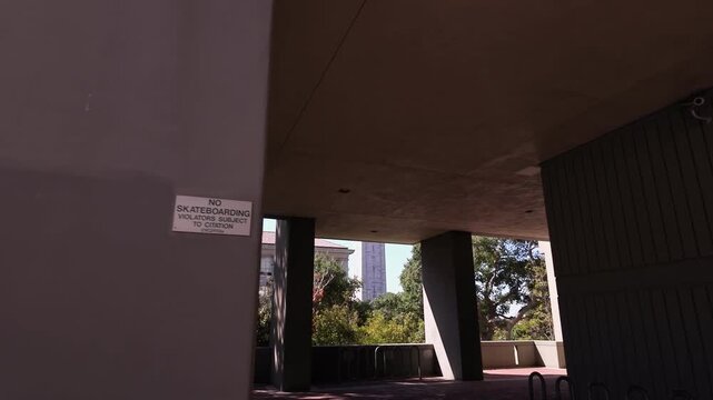 Personal perspective view walking under the concrete brutalist architecture of Evans Hall on the University of California, Berkeley campus, revealing the iconic Sather Tower on a sunny day