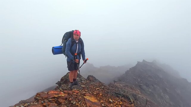 Female trekker climbs steep ridge, side wide view, carrying a loaded backpack through fog and loose rock in Svaneti, Georgia, illustrating expedition endurance, difficult terrain and travel challenges