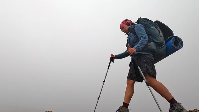 Female backpacker traverses rocky ridge, low side view, using trekking poles under a heavy load in foggy Svaneti, Georgia, supporting gear performance, balance training and expedition safety marketing