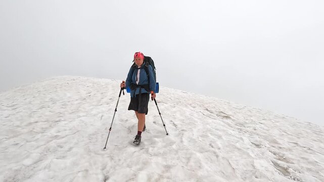 Female hiker crosses alpine snowfield, front wide view, using trekking poles in dense fog in Svaneti, Georgia, highlighting reduced visibility, seasonal route access and cold-weather travel safety