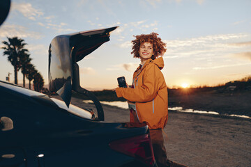 Fototapeta premium Woman lifestyle near open car trunk at sunset, curly hair portrait, holding smartphone, relaxed smile, warm golden light, casual jacket, outdoors travel mood, soft breeze, natural colors