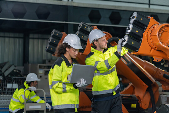 Engineers wearing hardhats using laptop computer to inspect and program robotic arm system in factory.