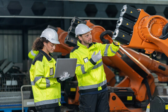 Engineers wearing hardhats using laptop computer to inspect and program robotic arm system in factory.