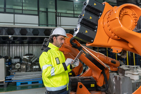 Technician wearing hardhat and safety vest holding tablet to inspect robotic arm in factory.
