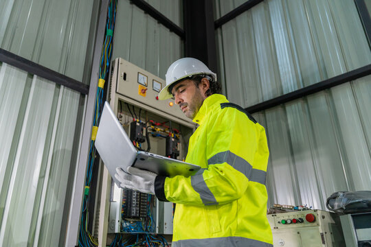 Technician wearing hardhat using laptop computer to inspect electrical control cabinet in factory.