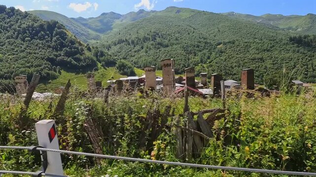 Traveler passing Svan towers in wide view beside a mountain village near Mestia, Georgia, presenting medieval defensive architecture for cultural tourism, heritage education and regional promotion