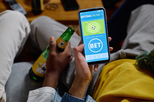 Diverse young adult male football fans holding beer bottle and using online betting app on smartphone. Concept of mobile gambling, sports entertainment and digital marketing, close up