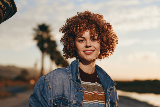 Cheerful woman lifestyle outdoors, curly hair, denim jacket, smiling in warm sunset light, relaxed posture, natural background, soft sky blur, candid portrait, golden tones, gentle wind