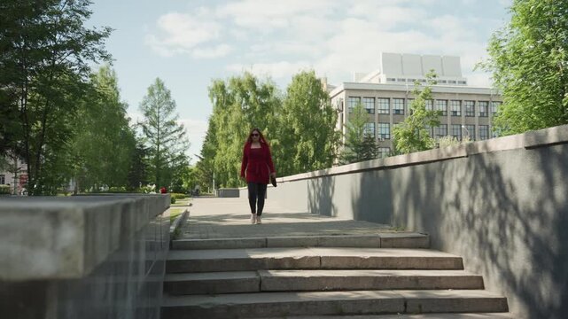 Woman in red coat walks down stone steps and pauses on landing holding small bag. scene features green trees lining pathways with buildings visible under blue skies creating outdoor urban