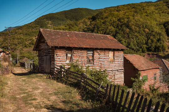 Old rustic mud brick and wooden house with tile roof beside a dirt path in Topli Do village, surrounded by green mountain hills at Stara Planina reserve in Serbia.