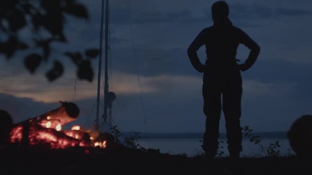 Sailing and camping in the wild. Woman sailor walks on the wild area with sailing boat on the background and bonfire on the foreground