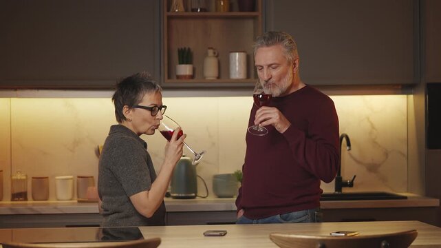 Cheerful married couple in their 50s drink wine in kitchen, enjoying evening