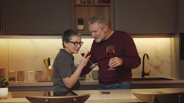 Happy mature couple drink wine and talk in kitchen, enjoying leisure together