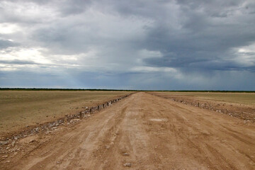 Eine gerade unbefestigte Straße führt durch eine weite, flache Landschaft  dunkle Wolken und sichtbare Regenfelder bestimmen den Himmel. © Jane Be. The Picture