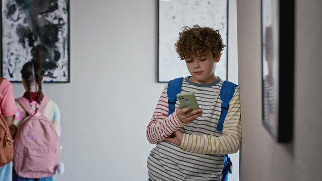 Caucasian child with backpack leaning against wall, using smartphone in art gallery while other kids look at paintings. Ideal for promoting educational apps or youth digital technology campaigns