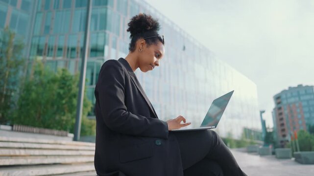 Businesswoman working with laptop in city street in daytime, side portrait. Female office worker in brake, online job mode out of office, it professional sending messages in work chat or writing code