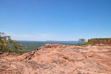 Eine flache rote Felsfläche im Vordergrund öffnet den Blick über grünes Buschland zu einer langen Fels- oder Hügellinie am Horizont. © Jane Be. The Picture