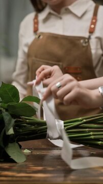 Vertical shot of Caucasian female florists tying ribbon around green rose stems on wooden table. Footage is ideal for small business marketing and floral design tutorials