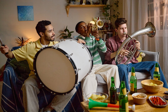 Diverse young adult male football fans playing musical instruments on sofa at home celebrating victory. Image suitable for sports entertainment and fan culture marketing campaigns