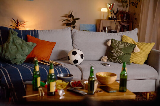 Empty living room with soccer ball on sofa and beer bottles on table after male football fans watching game, representing sports entertainment and home party commercial concepts