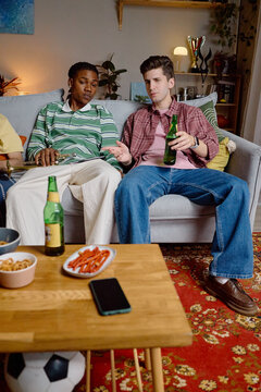 Vertical image of young adult African American and Caucasian male football fans sitting on sofa, drinking beer and discussing game, perfect for sports broadcast marketing
