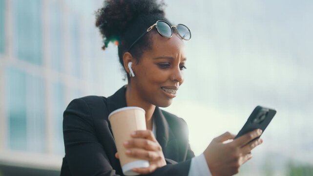 Businesswoman reading messages in smartphone while drinking coffee in park. Portrait of mixed race lady listening to music by earphones and choosing playlist in mobile phone, african american lady