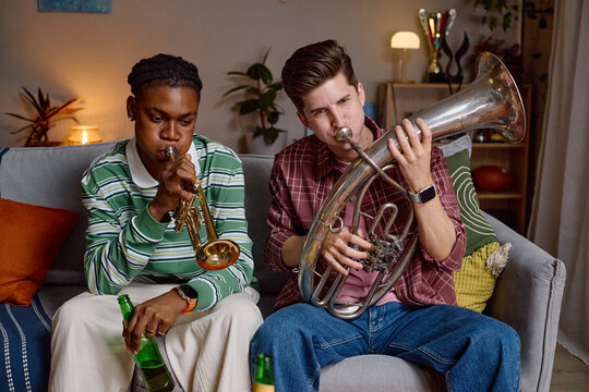 Diverse young adult male football fans sitting on sofa and playing wind instruments while celebrating victory. Image ideal for sports entertainment and fan culture marketing campaigns