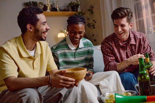 Diverse young adult male football fans sitting on sofa, laughing and holding snacks while watching game. Image suitable for sports broadcasting, entertainment, and social gathering campaigns