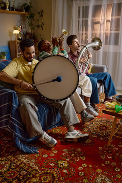 Vertical image of diverse young adult male football fans playing musical instruments at home while celebrating victory. Scene is suitable for sports marketing and fan culture campaigns