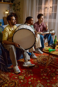 Vertical image of diverse young adult male football fans sitting on sofa, holding drum and trumpet while watching match. Scene represents sports entertainment and fan engagement