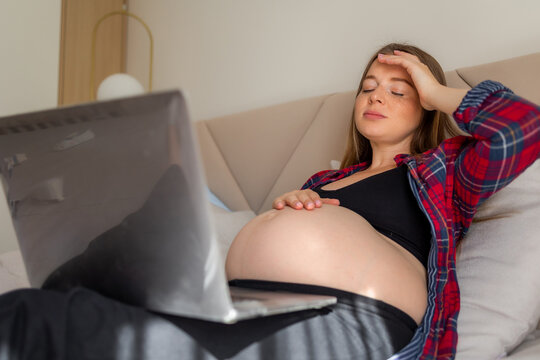 Pregnant woman reclining on a bed appearing tired balancing remote work and prenatal rest at home.