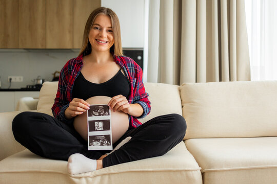 Pregnant woman at home smiling while holding ultrasound photos over her bare baby bump, expecting mother celebrating pregnancy