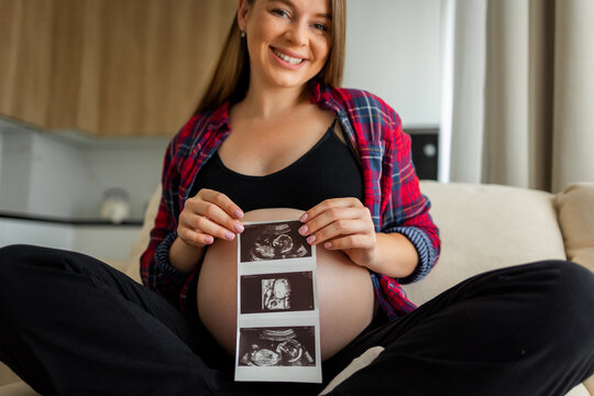 Smiling pregnant woman holding ultrasound photos over her bare belly celebrating upcoming baby and motherhood
