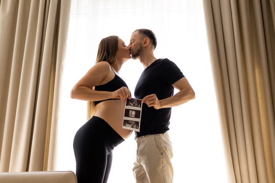 Pregnant couple kissing and holding ultrasound photo in a warm maternity announcement at home