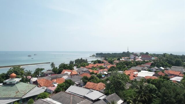 Aerial drone shot moving backward and upward revealing a coastal village with traditional houses, lush trees, a mosque, and the sea under a clear sky