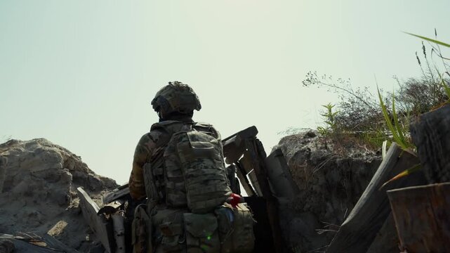 Back view of armed special forces soldier in full tactical gear and helmet running through narrow trench taking combat position among concrete fortifications and dry grass. Military warfare concept fo