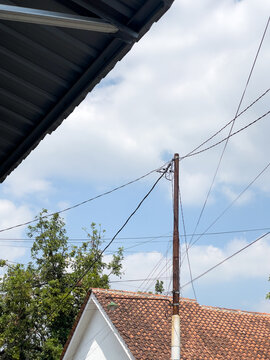 Utility pole with power lines over residential roofs. Daylight highlights textured clay tiles and metal. Intersecting wires create geometric depth. Ideal for infrastructure projects.