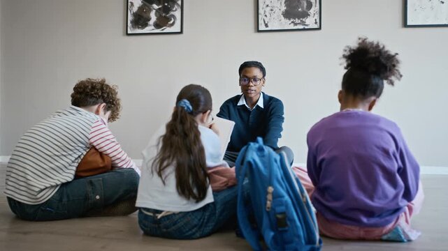 Young adult Black male teacher talking to diverse group of children sitting on floor in art gallery. Video is perfect for promoting museum education and interactive school field trips