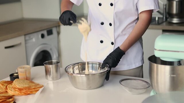 Professional female pastry chef whisking cream in a metal bowl while preparing a layered cake in a modern confectionery kitchen.