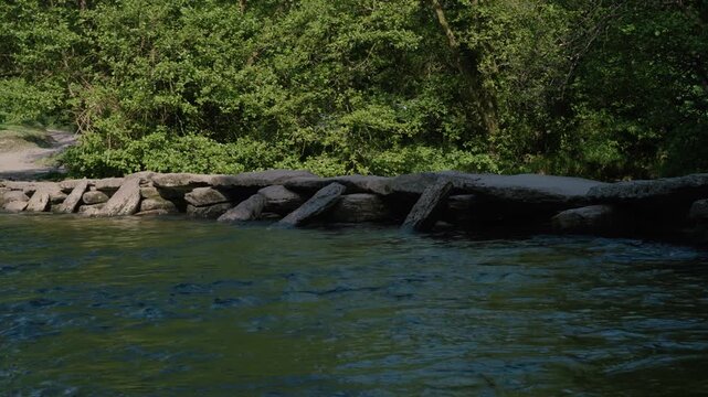 Tarr Steps in Exmoor National Park, focusing on the alignment of large stone slabs over the river. The composition includes soft ripples in the water, green foliage, and a tranquil atmosphere.