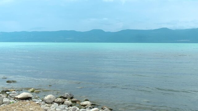 A scenic calm timelapse of Lake Iznik in Bursa Turkey featuring a rocky shoreline gentle water ripples and majestic distant mountains under an overcast sky