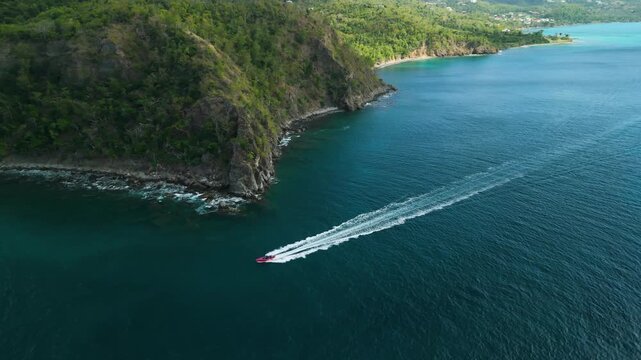 Aerial tracking follows red speedboat carving wake past forested cliffs and turquoise bay near Anse La Roche Carriacou