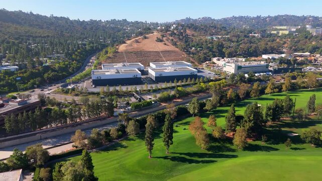 Aerial drone flies forward over rows of numbered soundstage buildings on the Universal Studios backlot, passing a manicured golf course with tall trees in autumn color, framed by hillside