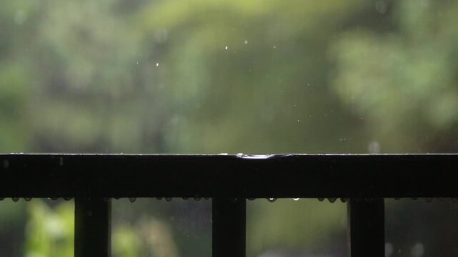 Slow motion footage of raindrops falling and splashing on a metal railing with blurred greenery in background.